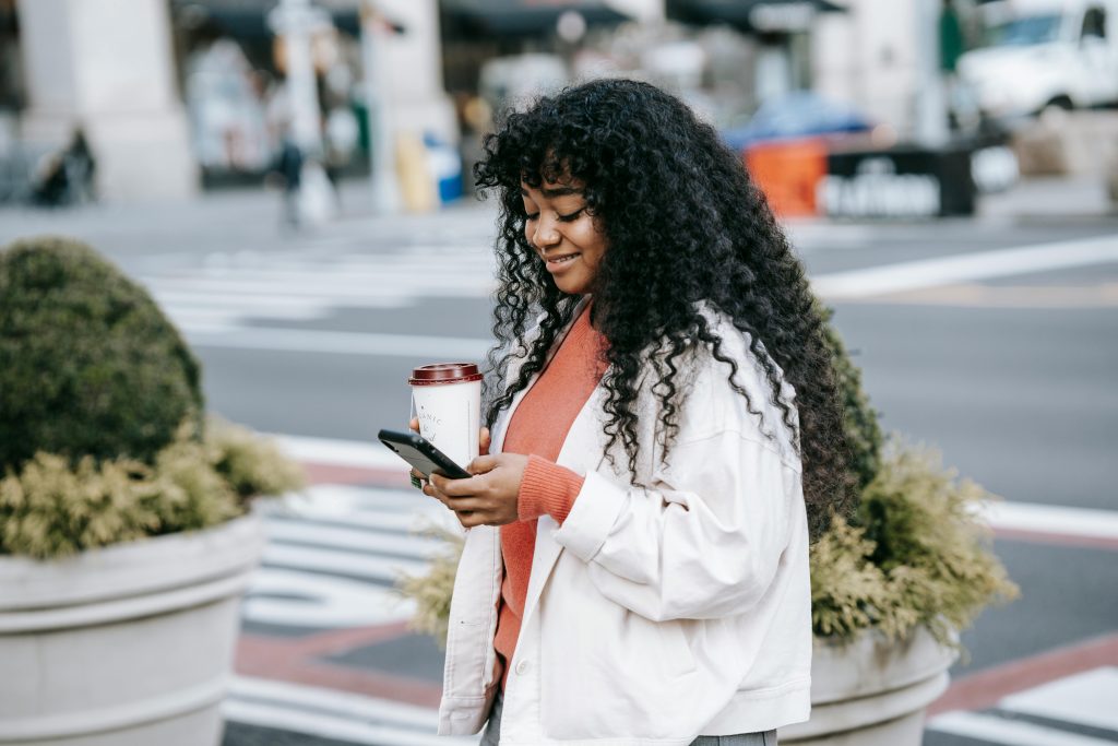 Cheerful adult African American female in white jacket surfing mobile phone while standing with takeaway coffee on modern city street